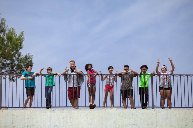 Group of children standing together on a concrete bridge railing, smiling and posing for the camera on a sunny day