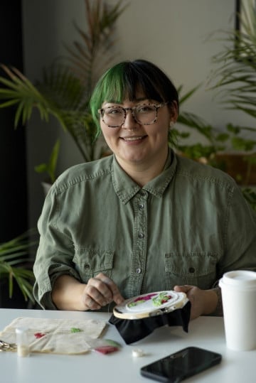 Person with green-tinted hair and glasses wearing a sage green shirt, smiling at camera while holding a plate of food at a table with potted plants in background