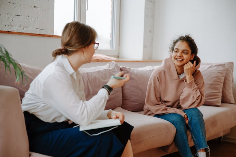 Female therapist taking notes during counseling session with smiling young woman on beige couch in bright office